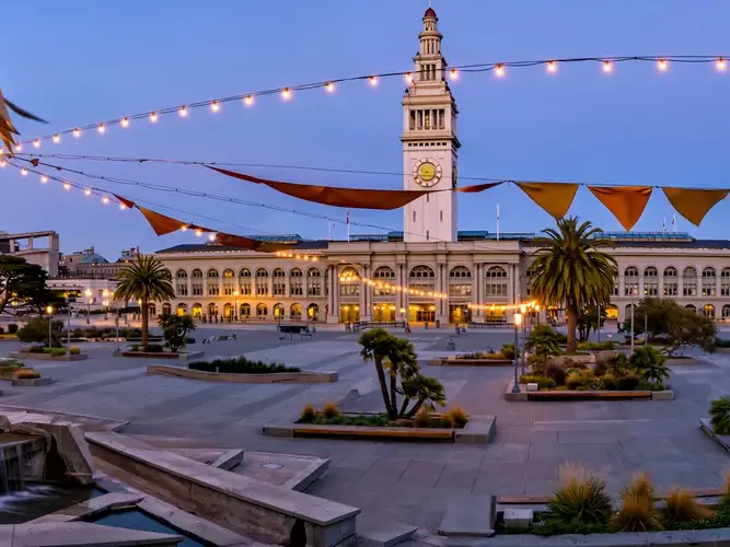 A large public building with a clock tower, surrounded by a plaza with palm trees and string lights.